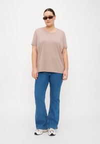 Voľný béžový tričko s krátkymi rukávmi v kombinácii s modrými zvonovými džínami a bielymi teniskami. Hnedé slnečné okuliare dokončujú outfit.