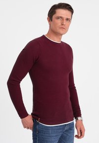 Burgundy long-sleeve shirt with a textured weave, featuring a contrasting white neckline. Paired with blue jeans.