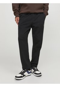 Osoba v černých teplácích se založenýma rukama v kapsách a v černobílomodrých teniskách stojící před jednobarevným světlým pozadím.