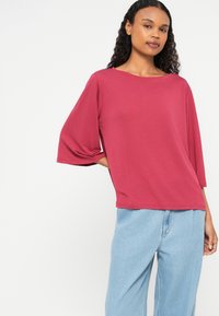 Maroon relaxed-fit top with wide sleeves, a rounded neckline, and a smooth texture. Paired with light blue high-waisted jeans.