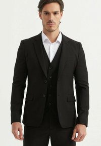 Costume noir sur mesure avec revers cranté, accompagné d'une chemise blanche. Les boutons sur la veste et le gilet créent une apparence élégante et formelle.