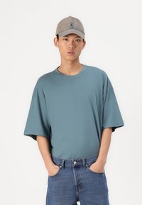 Camiseta de gran tamaño en azul claro, con mangas cortas, confeccionada en material de algodón y con un cuello redondo. Combinada con pantalones cortos de mezclilla azules y una gorra gris claro.