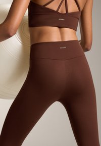 Hnedé športové legíny a zodpovedajúci crop top s elegantným dizajnom, obsahujúci vysoký pás a logo na chrbte. Hladká, elastická tkanina.