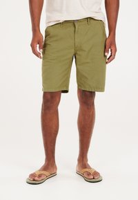 Olivgrüne Baumwollshorts mit flacher Vorderseite, zwei Seitentaschen und Knopfverschluss. Getragen mit beigen Flip-Flops auf baren Füßen.