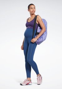 Top desportivo roxo, leggings azul-marinho e sapatos atléticos cor-de-rosa. A carregar uma mala lilás clara com um padrão geométrico. Materiais lisos e elásticos.