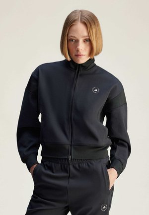 Black zippered jacket made of smooth fabric, featuring a high collar, side pockets, and a logo on the left chest. Paired with matching trousers.