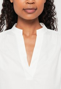 Bluză albă din bumbac cu guler chinezesc, având un decolteu în formă de V și o textură moale, cu mâneci scurte puff.