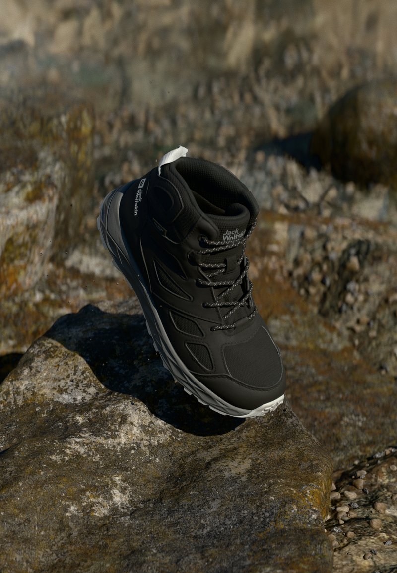 Bota de caminhada preta com um parte superior texturizada, detalhes em cinza e uma sola branca. Apresenta atacadores e um colarinho almofadado, repousando numa superfície rochosa.