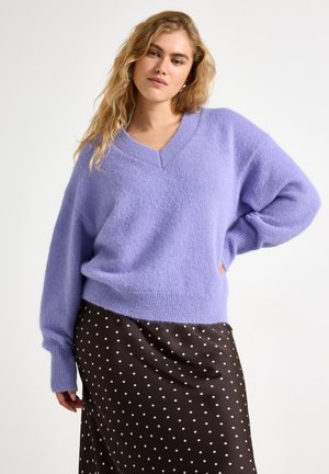 Kvinde iført en lavendelfarvet, blød V-hals sweater og et sort skørt med hvide polkaprikker, poserer med den ene hånd på hoften mod en hvid baggrund.