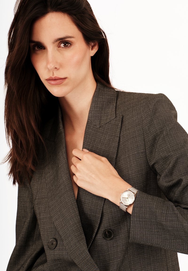 Montre argentée avec un cadran rond et un bracelet en mailles, portée au poignet. Le modèle porte un blazer gris foncé, texturé, avec de larges revers.