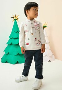 Weißer Strickpullover mit festlicher Stickerei und Aufnähern, kombiniert mit marineblauen Hosen und weißen Sneakers. Im Hintergrund grüne und weiße Papier-Weihnachtsbäume.