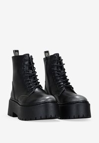 Botas de tobillo de cuero negro con suela de plataforma gruesa, punta redonda y cordones delanteros. Presentan costuras decorativas y tiradores en los talones.