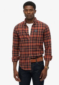 Red and navy plaid flannel shirt with long sleeves, button-down collar, and a logo patch on the chest. Worn with dark jeans and a brown belt.