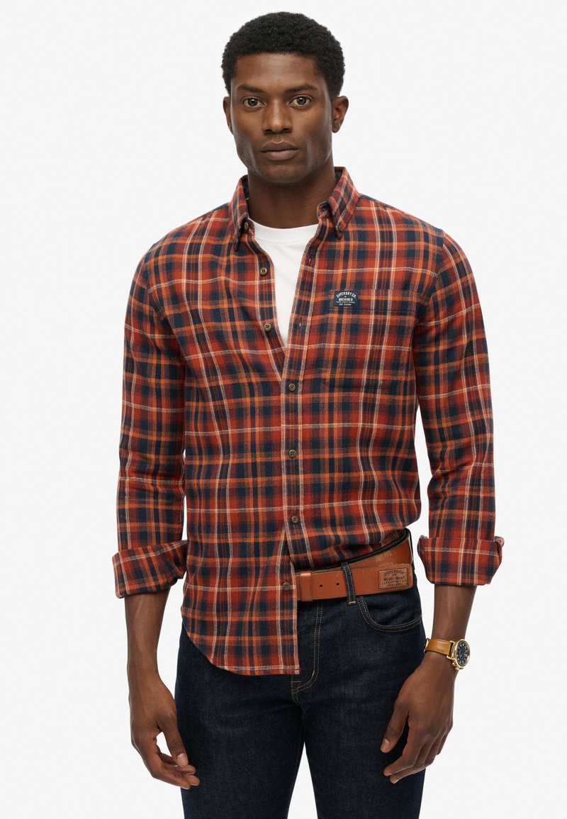 Red and navy plaid flannel shirt with long sleeves, button-down collar, and a logo patch on the chest. Worn with dark jeans and a brown belt.