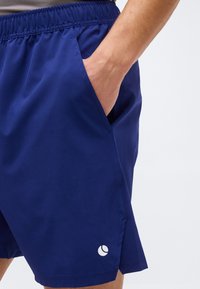 Άνδρας που φοράει μπλε αθλητικά σορτς με ελαστική μέση και το χέρι στην πλαϊνή τσέπη, με ορατό μικρό λευκό λογότυπο κοντά στο κάτω μέρος.