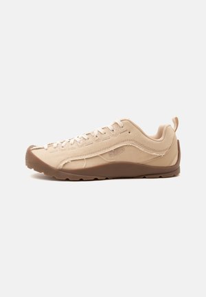 Beige lavtop sneaker med hvide snørebånd, brun sål, lærredsupper og trækflap ved hælen, vist fra ydersiden mod en ensfarvet baggrund.