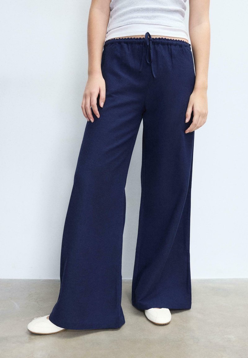 Personne portant un pantalon large bleu marine avec taille à cordon et des chaussures blanches sans lacets, sur un fond clair uni.