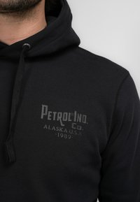 Sudadera con capucha negra de algodón. Presenta una impresión texturizada del logo que dice "Petrol Ind. Co. Alaska U.S.A. 1989" en el pecho.