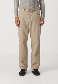 Man med beige raka byxor, grå ribbad long-sleeve tröja och svarta sneakers med vita ränder, stående framför en vit bakgrund.