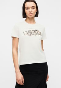 Νεαρή γυναίκα με ίσια σκούρα μαλλιά, φορώντας ένα λευκό μπλουζάκι με το κείμενο «VINTAGE» και λεοπάρ μοτίβο, συνδυασμένο με μια μαύρη φούστα.