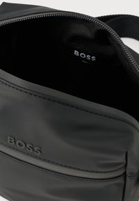 Μαύρη nylon τσάντα χιαστί με φερμουάρ, ομαλή υφή, ανάγλυφο λογότυπο και εσωτερική ετικέτα με το όνομα της μάρκας.