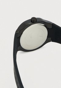 Musta Guess-rannekello, jonka takakansi on hopeanväristä ruostumatonta terästä ja jossa on teksturoitu musta ranneke.