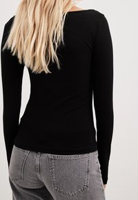 Schwarz geripptes Langarmoberteil mit tailliertem Design. Von hinten gesehen hat es einen runden Halsauschnitt und wird mit grauen hochtaillierten Jeans kombiniert.