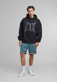 Czarna bluza z kapturem z dużym szarym logo "K1X", w połączeniu z turkusowymi szortami z siateczki z czarnymi i białymi paskami. Białe sneakersy.