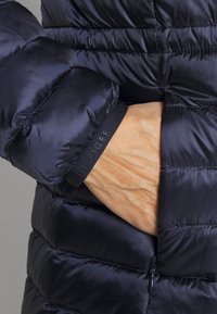 Laivastonsininen toppatakki, jossa on tikattu rakenne ja joustavat hihansuut. Varustettu mustalla brändätyllä nauhalla ja sivutaskulla. Sileä, heijastava materiaali.