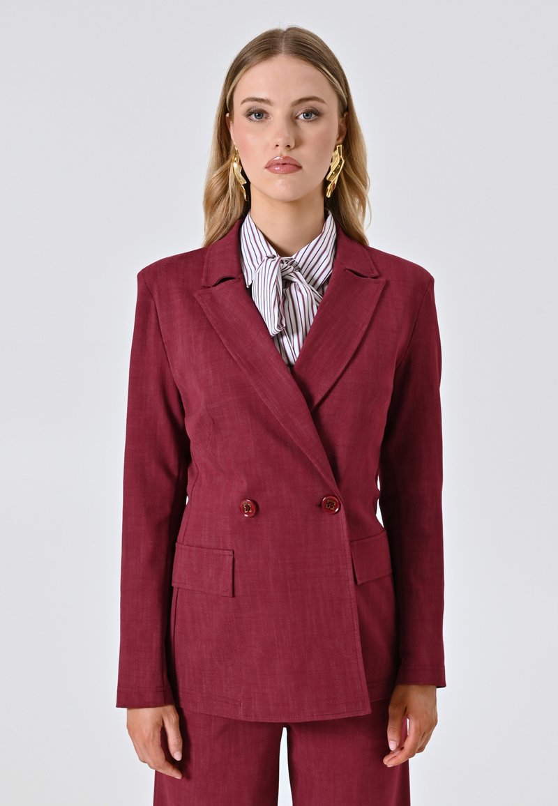 Bordeauxfarbener, doppelt-breaster Blazer mit Reverskragen, zwei Fronttaschen und dekorativen Knöpfen. Kombiniert mit einer gestreiften Bluse mit Bindegürtel.