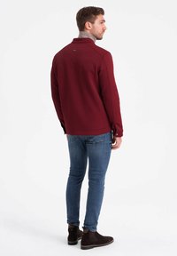 Mörkröd zip-up jacka med ett texturerat mönster, hög krage och sydda detaljer, tillsammans med blå jeans och mörkbruna stövlar.