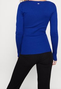Royal blue fitted sweater with long sleeves and ribbed accents at the waist. Smooth texture, round neckline, features a small tag detail at the back.