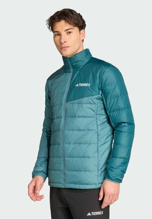 Uomo che indossa una giacca a piumino Terrex color turchese e verde acqua e pantaloni Terrex neri, in piedi davanti a uno sfondo chiaro e semplice.