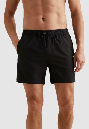 Schlanker männlicher Oberkörper und Beine, die schwarze Badeshorts mit Kordelzug tragen, mit den Händen in den Taschen, vor einem einfarbigen hellen Hintergrund.