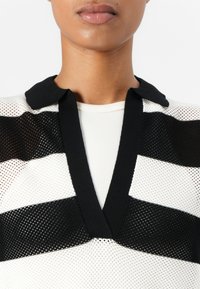 Svart og hvit stripete strikket topp med mesh tekstur, med V-hals og solid svart krage. Designet er preget av dristige mønstre.