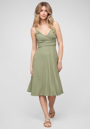 Vrouw met lang blond haar die een mouwloze lichtgroene midi-jurk en beige sandalen draagt, loopt vooruit tegen een egale lichte achtergrond.