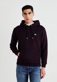 Felpa con cappuccio di colore viola scuro realizzata in misto cotone, con tasca frontale, cappuccio con lacci e piccolo logo bianco sul petto. Indossata con jeans di denim.