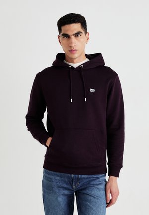 Felpa con cappuccio di colore viola scuro realizzata in misto cotone, con tasca frontale, cappuccio con lacci e piccolo logo bianco sul petto. Indossata con jeans di denim.