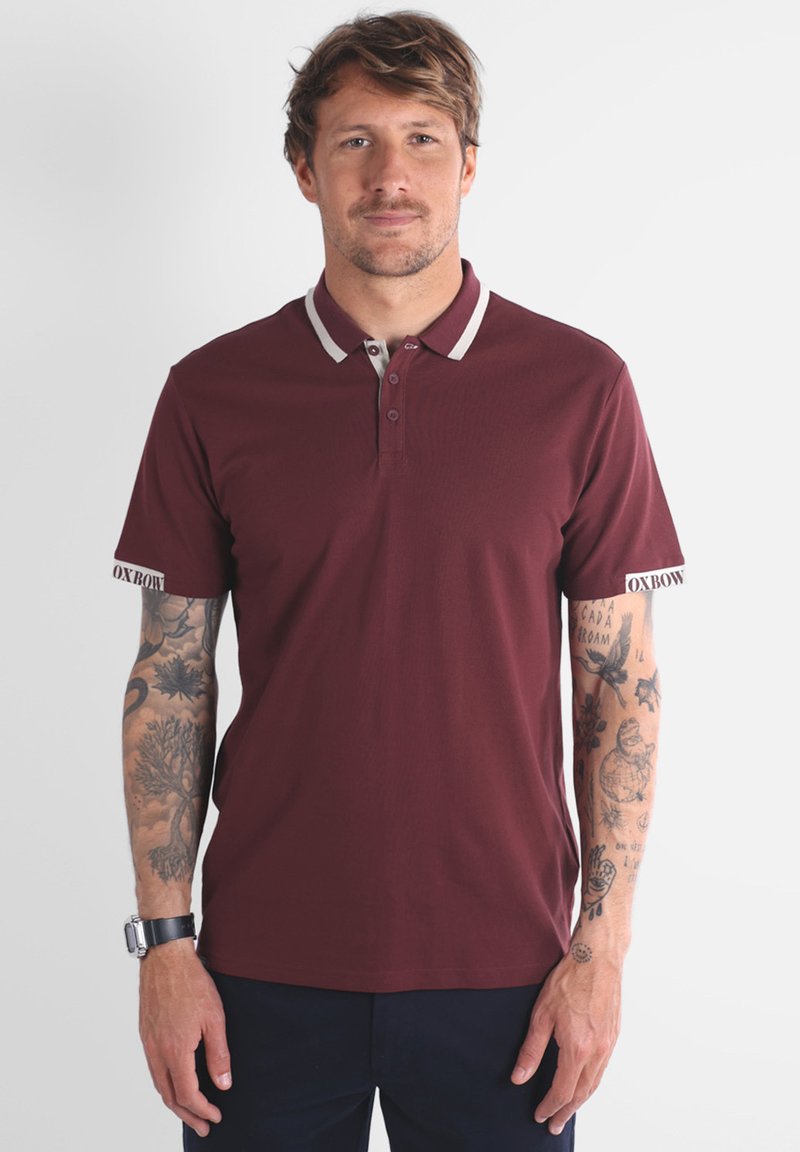Homme avec des cheveux châtain clair portant un polo maroon avec des bordures blanches et des tatouages couvrant les deux avant-bras, se tenant devant un fond blanc.