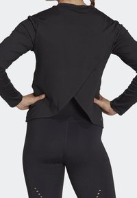 Svart långärmad topp med överlappande design bak, mjuk textur och figurnära silhuett. Matcher med svarta leggings med accentuerade utskärningar.