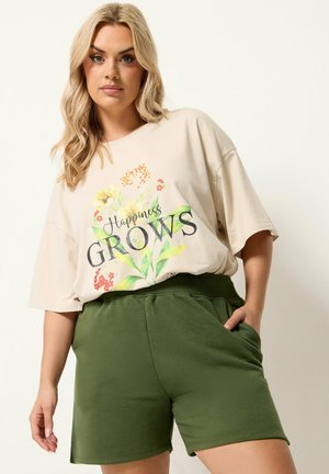 Kobieta nosząca beżowy, oversize T-shirt z kwiatowym nadrukiem "Happiness Grows", wpuszczony w zielone szorty z elastycznym pasem, pozująca z jedną ręką w kieszeni.