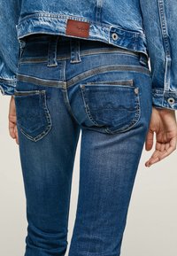 Tmavě modré skinny džínsy s hladkým povrchem, mají zadní kapsy a kontrastní stehy. Na pase je viditelný kožený štítek.