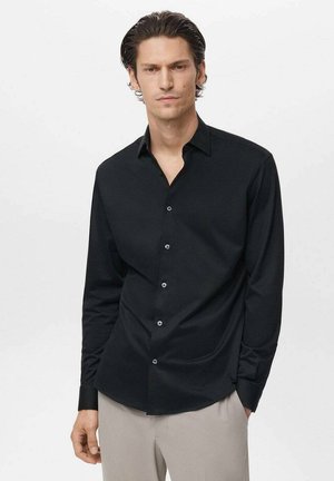 Hombre vestido con una camisa de botones de manga larga negra y pantalones beige claros, de pie con una mano en el bolsillo frente a un fondo sencillo.