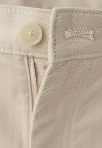 Pantalones beige con un patrón de espiga, un cierre con un gran botón y una cremallera oculta en la parte delantera. Tejido resistente con textura suave.