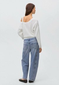 En kvinna med långt hår bär en vit off-shoulder tröja, blå jeans och svarta ballerinatöj, sedd bakifrån mot en ljus bakgrund.