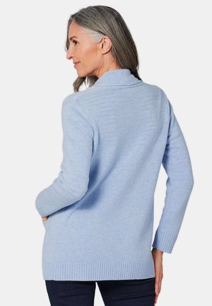 Cardigan bleu clair en tricot doux, avec un col châle et des poignets et un ourlet côtelés. Deux poches latérales sont visibles.