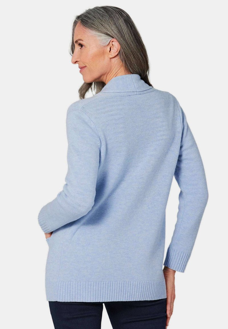 Cardigan bleu clair en tricot doux, avec un col châle et des poignets et un ourlet côtelés. Deux poches latérales sont visibles.