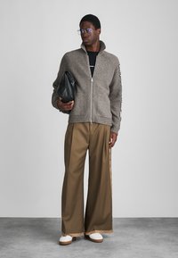 Pletený šedý svetr na zip, široké béžové kalhoty s bočními pruhy, bílé tenisky, držící černou tašku. Minimalistický, moderní design.