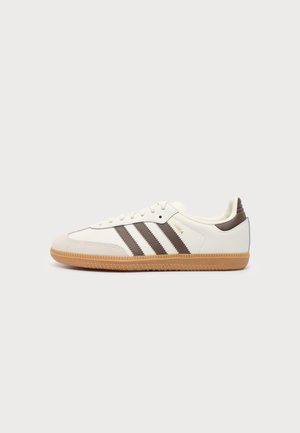 Baskets blanches avec bandes Adidas marron, semelle en gomme, profil bas et texte doré "SAMBA" sur le côté, présentées sur un fond blanc uni.