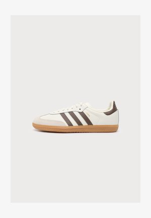 Λευκό αθλητικό παπούτσι με καφέ ρίγες Adidas, καουτσούκ σόλα, χαμηλό προφίλ και χρυσή επιγραφή "SAMBA" στο πλάι, σε απλό λευκό φόντο.